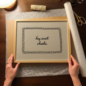 May include: A framed print with the text "hey sweet cheeks" in a decorative font. The print is in a light wood frame, held by hands. Bubble wrap, tape, scissors, and a cup of coffee are on the table.
