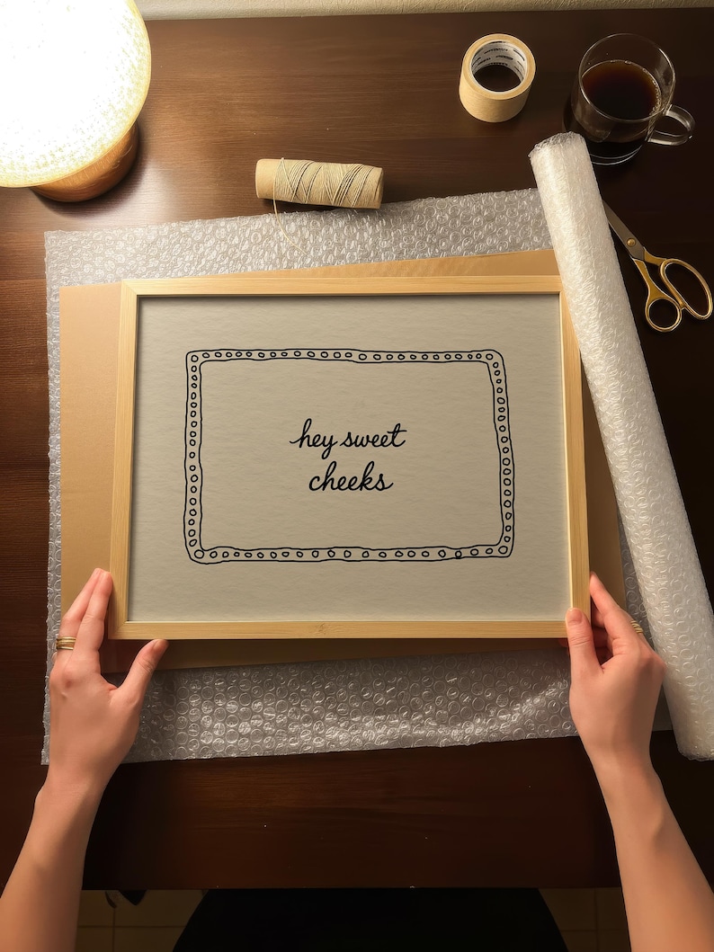 May include: A framed print with the text "hey sweet cheeks" in a decorative font. The print is in a light wood frame, held by hands. Bubble wrap, tape, scissors, and a cup of coffee are on the table.