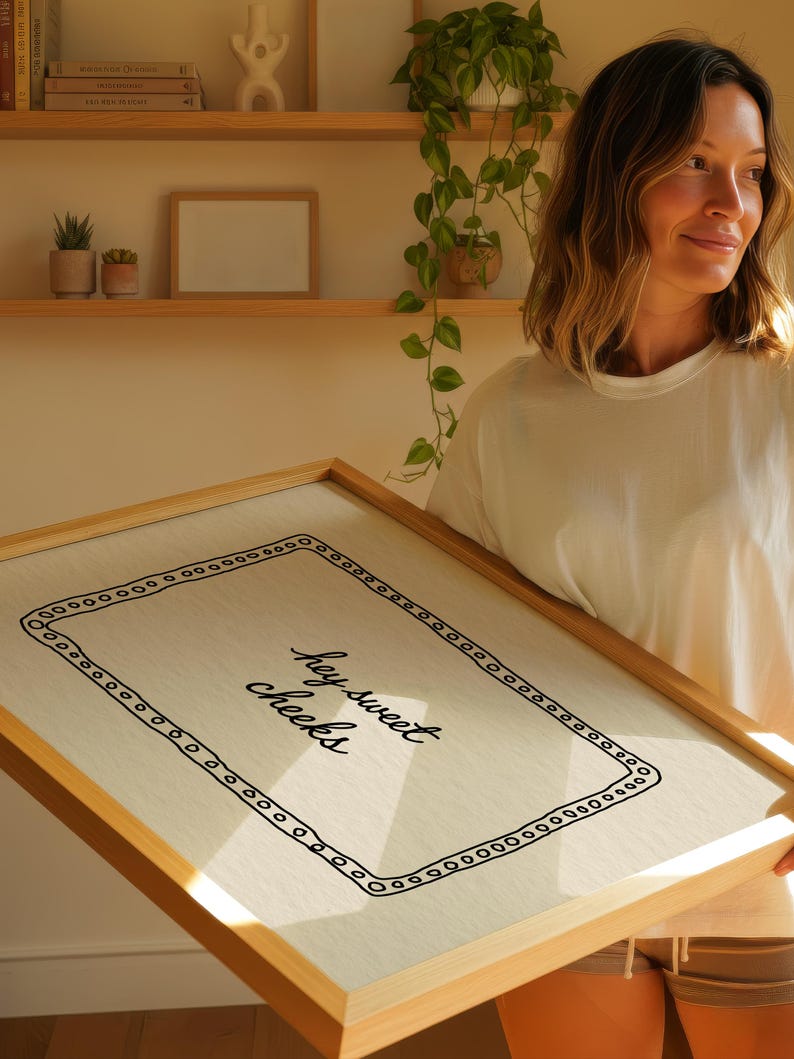 May include: A framed canvas print with the text "hey sweet cheeks" in a handwritten style. The print has a light-colored background with a black border of small circles. The frame is made of light-colored wood.