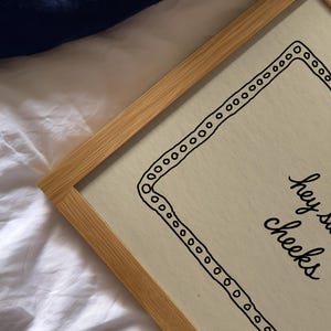 May include: A framed print with a light wood frame. The print has a cream-colored background with a black border of circles and the words "hey sweet cheeks" in black script.
