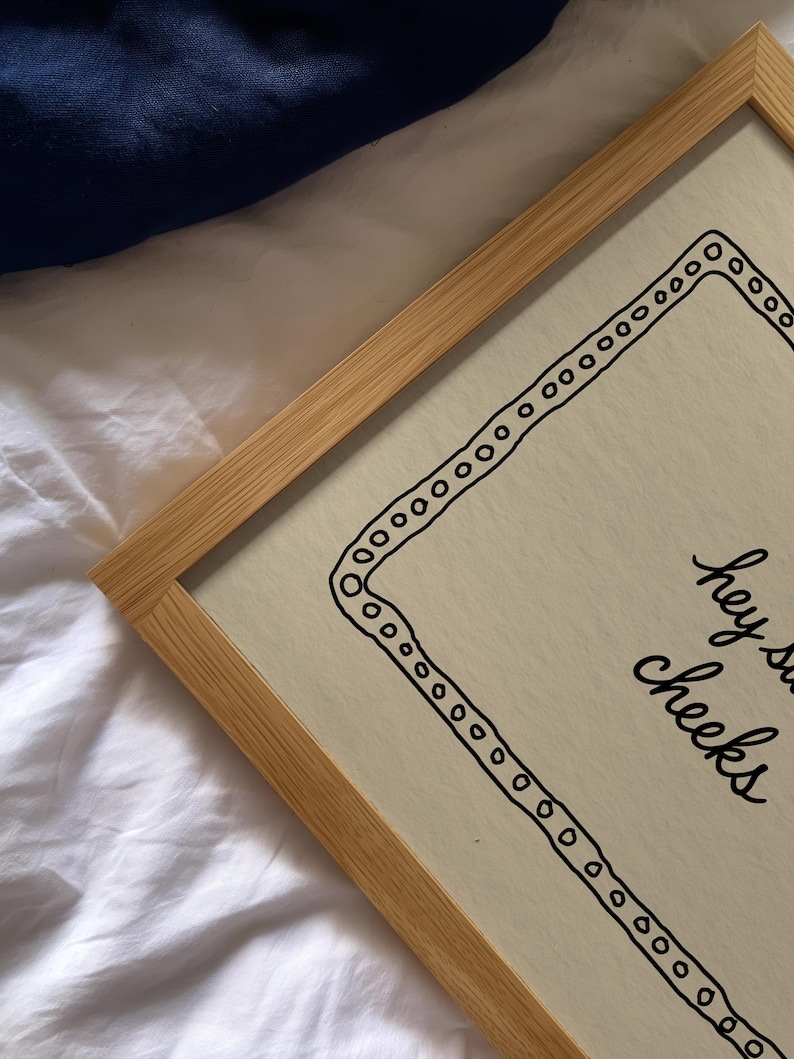 May include: A framed print with a light wood frame. The print has a cream-colored background with a black border of circles and the words "hey sweet cheeks" in black script.