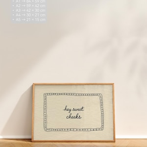 May include: A framed print with the text "hey sweet cheeks" in a handwritten style. The print has a light beige background with a black border of small circles and a wooden frame. The print sizes are listed in centimeters.