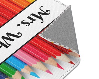 May include: A rectangular rug featuring a colorful pencil design. The pencils are arranged in a row, with their tips pointing towards the right. The rug has a white border with the text "Mrs. W" in black.