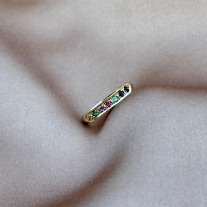 Puede incluir: Un anillo dorado con una fila de piedras preciosas de colores. Las piedras preciosas incluyen piedras verdes, moradas, rojas y azules. El anillo está sobre un fondo suave y de tono neutro.