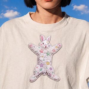 May include: An off-white t-shirt featuring a large embroidered patch of a white rabbit with its limbs spread wide. The rabbit is decorated with pastel-colored embroidered stars, small circles, and flower shapes in pink, green, yellow, and blue.