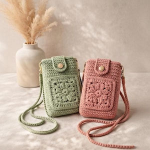 Op de afbeelding: Twee gehaakte telefoonzakjes in saliegroen en oudroze. Elke tas heeft een bloemenmotief op de voorkant, een houten knoopsluiting en een lange, gevlochten band. De tassen zijn ongeveer 18 cm hoog en 10 cm breed.