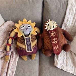 May include: Two plush toys featuring stylised characters. The left plush has a yellow sun-shaped head, a grey face, and a purple patterned body with a gold-trimmed tag. The right plush is brown with a white sun-shaped head and a red vertical tag.