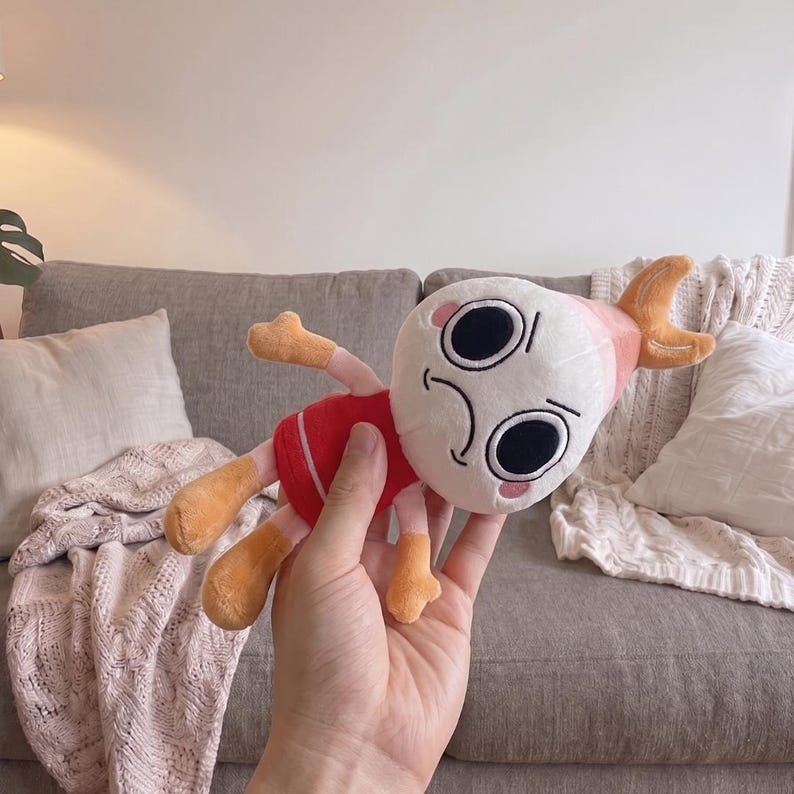 Puede incluir: Un peluche con cabeza blanca redondeada, grandes ojos negros circulares, una peque&ntilde;a boca curvada y mejillas rosadas. El personaje tiene un torso rojo, extremidades de color naranja claro y una punta de color naranja claro en la cabeza.