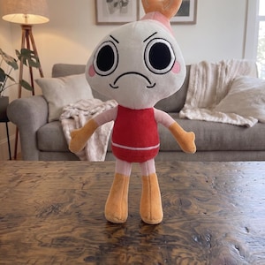 Puede incluir: Un peluche con una cabeza blanca grande de expresi&oacute;n enfadada, ojos negros circulares y mejillas rosadas. El personaje tiene un torso rojo, brazos de color rosa claro y manos y pies naranjas. Una peque&ntilde;a cola de camar&oacute;n naranja corona la cabeza.