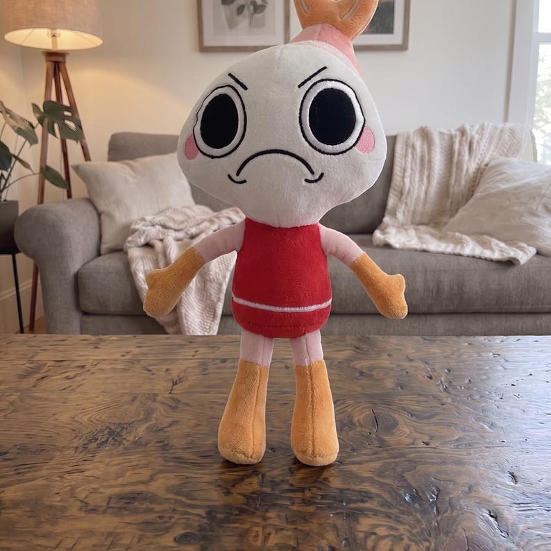 Puede incluir: Un peluche con una cabeza blanca grande de expresi&oacute;n enfadada, ojos negros circulares y mejillas rosadas. El personaje tiene un torso rojo, brazos de color rosa claro y manos y pies naranjas. Una peque&ntilde;a cola de camar&oacute;n naranja corona la cabeza.