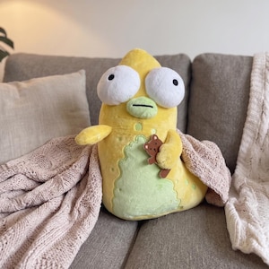 Può includere: Un peluche giallo a forma di creatura dei cartoni animati con grandi occhi bianchi e una pancia verde. Tiene in mano un piccolo orsacchiotto marrone ed è drappeggiato con una coperta rosa. Il giocattolo è su un divano grigio.