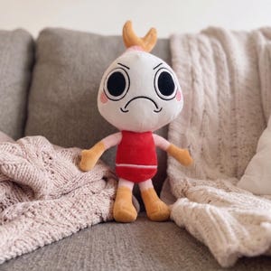 Puede incluir: Un peluche con cabeza redonda blanca, grandes ojos negros, boca fruncida y mejillas rosadas. Tiene una peque&ntilde;a cola de camar&oacute;n naranja en la parte superior de la cabeza, un torso rojo, brazos de color rosa claro y piernas amarillas.