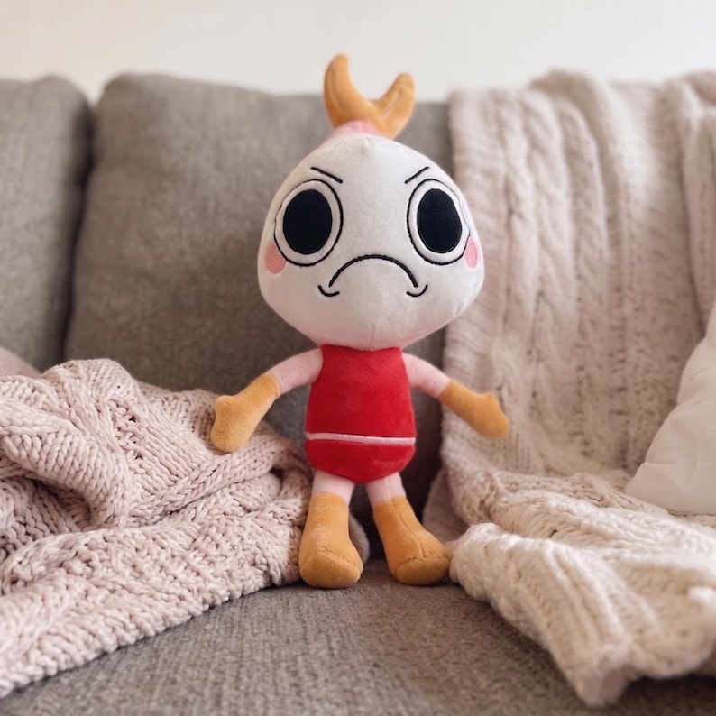 Puede incluir: Un peluche con cabeza redonda blanca, grandes ojos negros, boca fruncida y mejillas rosadas. Tiene una peque&ntilde;a cola de camar&oacute;n naranja en la parte superior de la cabeza, un torso rojo, brazos de color rosa claro y piernas amarillas.