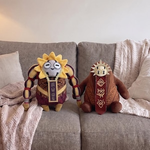May include: Two plush toys featuring stylized sun-like headpieces. The left toy has a yellow sun crown, a grey face, and a patterned maroon body with a rectangular pendant. The right toy is brown with a white sun crown and a long red necktie.