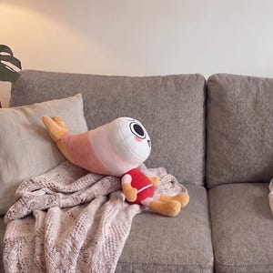 Puede incluir: Un peluche con forma de camar&oacute;n que tiene una cabeza blanca, ojos negros grandes y un cuerpo rosado. El personaje viste una camiseta roja, pantalones cortos de color rosa claro y tiene extremidades color canela. La cola es de color naranja.