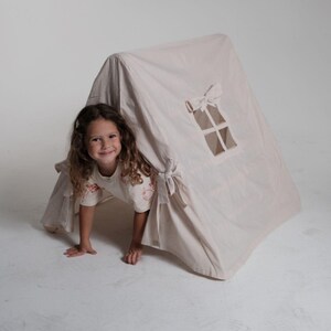 May include: A beige playhouse tent with a window and bow accents. A child is peeking out from under the tent, smiling. The tent is made of a soft fabric and has a simple, classic design, perfect for indoor or outdoor play.