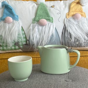 Puede incluir: Una tetera de cerámica verde claro con un infusor y asa de acero inoxidable, junto a una taza a juego. La tetera y la taza están sobre una superficie gris, con un cojín decorativo con diseños de gnomos en el fondo.