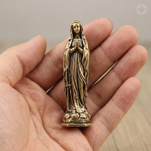 Puede incluir: Peque&ntilde;a estatua de la Virgen Mar&iacute;a en color bronce, con las manos juntas en oraci&oacute;n. La estatua presenta una detallada indumentaria y un rosario. La figura est&aacute; sobre una peque&ntilde;a base.
