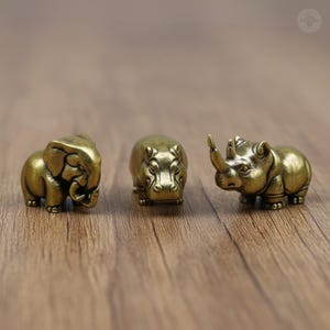 May include: Three small, bronze-coloured animal figurines: an elephant, a hippopotamus, and a rhinoceros. The elephant has detailed trunk and ear features. The hippo is compact, and the rhino has a prominent horn. All are on a wooden surface.
