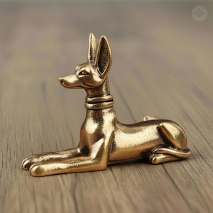 Può includere: Una statua color bronzo di un cane, forse Anubi, in posizione seduta. Il cane ha orecchie lunghe e dritte e un collare intorno al collo. La statua è dettagliata e sembra essere di metallo.