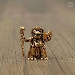 May include: A bronze frog figurine, dressed as a wizard, holding a staff and a book. The frog wears a hat and cape, with intricate details on the staff and clothing. The figurine is set against a wooden background.