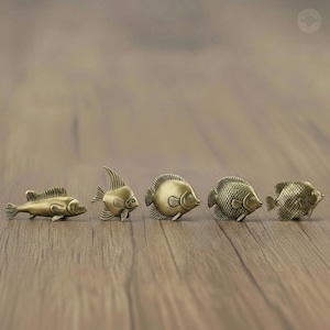 Könnte beinhalten: Fünf Bronzefischfiguren in verschiedenen Größen sind auf einer Holzoberfläche angeordnet. Jeder Fisch hat detaillierte Schuppen und Flossen mit einem Vintage-Look. Die Fische sind in einer Reihe angeordnet und präsentieren eine Sammlung von Wasserkörperdekorationen.