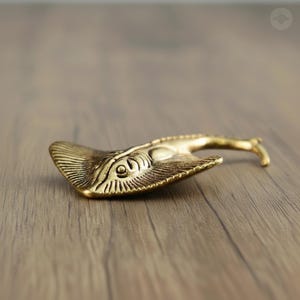 Puede incluir: Un abridor de botellas de latón con forma de manta raya, con grabados detallados. El abridor tiene un mango curvo y está sobre una superficie de madera. El diseño de la manta raya incluye detalles de ojos y aletas.