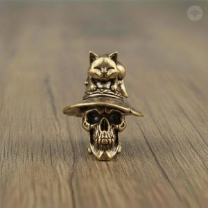 May include: A bronze-colored skull figurine wearing a witch hat, with a smiling cat perched on top. The skull has detailed features, and the hat has a wide brim. The cat adds a whimsical touch to the gothic design.