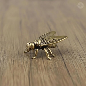 May include: A detailed bronze-colored metal fly figurine with intricate wing and body details. The insect is posed on a wooden surface, showcasing its legs and antennae. The sculpture is a decorative item.