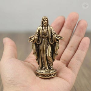Puede incluir: Una estatua de bronce de Jesucristo con los brazos extendidos, de pie sobre una base decorativa. La estatua presenta t&uacute;nicas detalladas y un emblema de coraz&oacute;n en el pecho. La figura se sostiene en una mano abierta.