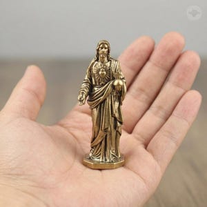 Puede incluir: Una peque&ntilde;a estatua de color bronce de una figura religiosa, sostenida en una mano. La figura est&aacute; representada con t&uacute;nicas fluidas, con un rostro detallado y un emblema de coraz&oacute;n en el pecho. La estatua mide aproximadamente 5 cm de altura.