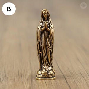 Puede incluir: Una peque&ntilde;a estatua de color bronce de una mujer con una t&uacute;nica fluida, con las manos juntas en oraci&oacute;n. La estatua tiene un dise&ntilde;o detallado, incluyendo un rosario y una base detallada. La figura est&aacute; sobre una base detallada.