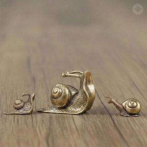 May include: Three brass snail figurines of varying sizes arranged in a row. Each snail features a textured shell and body with a metallic gold finish. The central snail is the largest, while the two smaller snails are positioned on either side.