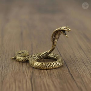 Puede incluir: Figurilla detallada de cobra de latón con la boca abierta, lista para atacar. La serpiente está enrollada, mostrando escamas texturizadas y una capucha levantada. La figurilla es de color dorado y está sobre una superficie de madera.