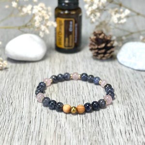 Könnte beinhalten: Ein Armband mit blauen, rosa und Holzperlen, akzentuiert durch goldfarbene Perlen. Im Hintergrund sind eine Flasche ätherisches Öl und ein Tannenzapfen zu sehen.
