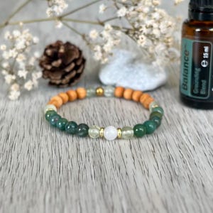 Può includere: Un braccialetto di perline verdi, bianche e di legno, con un accento dorato. Il braccialetto è esposto con un flacone di olio essenziale ed elementi decorativi.