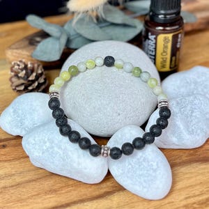 Könnte beinhalten: Ein Armband mit schwarzen Lavasteinperlen und hellgrünen und weißen Edelsteinperlen. Silberfarbene Metallabstandshalter akzentuieren das Armband. Im Hintergrund befindet sich eine Flasche ätherischen Öls mit dem Text "Wild Orange".