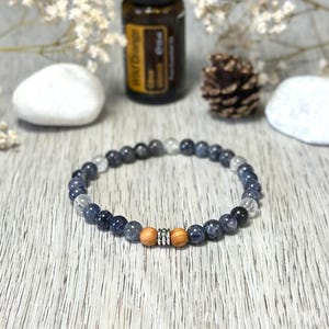 Könnte beinhalten: Ein Armband mit dunkelblauen und klaren Perlen, akzentuiert durch zwei Holzperlen und einen silbernen Abstandshalter. Eine Flasche ätherisches Öl und ein Tannenzapfen befinden sich im Hintergrund. Das Armband wird auf einer hellen Holzmaserung präsentiert.