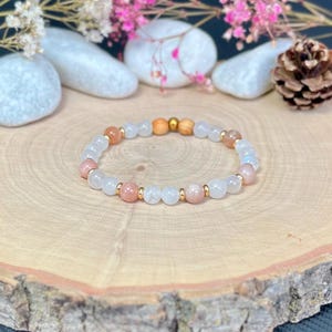 Può includere: Un braccialetto di perline bianche, color pesca e di legno, intervallate da piccoli elementi dorati. Il braccialetto è presentato su una superficie di legno, con fiori e pietre decorative sullo sfondo.