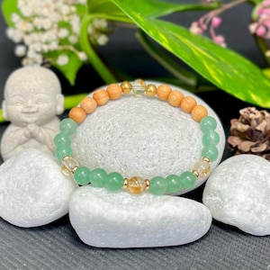 Könnte beinhalten: Ein Armband mit Perlen in Grün, Hellbraun und Gold. Das Armband wird auf einem weißen Stein präsentiert, mit einer kleinen Statue und Blumen im Hintergrund. Das Armband besteht aus natürlichen Materialien.