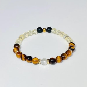 Può includere: Un braccialetto di perline con una combinazione di pietre preziose naturali. Il braccialetto include occhio di tigre, citrino, quarzo trasparente e perline di lava nera. Distanziatori color oro sono intercalati nel design. Il braccialetto è infilato su un cordoncino elastico.