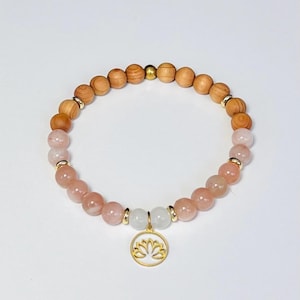 Può includere: Un braccialetto di perline con perline di legno marrone chiaro, perline di pietre preziose rosa e bianche e dettagli dorati. Un ciondolo dorato con un disegno di fiore di loto bianco pende dal braccialetto. Sfondo bianco.