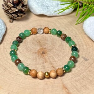 Puede incluir: Una pulsera de cuentas con cuentas verdes, marrones y de color madera clara. La pulsera incluye detalles dorados y una cuenta de cristal transparente. Se muestra sobre una superficie de madera.