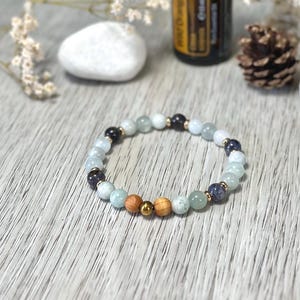 Könnte beinhalten: Ein Armband mit hellblauen, schwarzen und Holzperlen. Goldfarbene Abstandshalter akzentuieren das Design. Im Hintergrund eine Flasche, ein weißer Stein und Trockenblumen.
