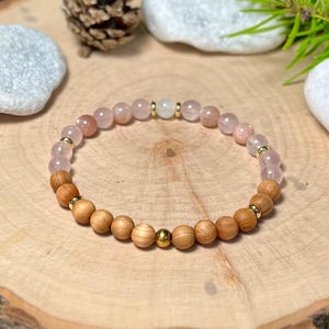 Könnte beinhalten: Ein Perlenarmband mit Rosenquarz- und Holzperlen, unterbrochen von goldfarbenen Akzenten. Das Armband wird auf einer Holzoberfläche präsentiert, die seine natürlichen Töne und Texturen hervorhebt. Es ist ein Schmuckstück.