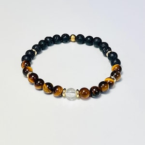 Op de afbeelding: Een kralenarmband met zwarte lavasteen kralen, tijgeroog kralen en een heldere kwartskraal. Goudkleurige afstandhouders accentueren de natuurstenen kralen. De armband is ontworpen voor dagelijks gebruik.
