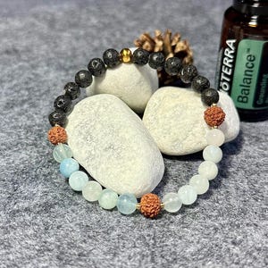 Könnte beinhalten: Ein Perlenarmband mit schwarzen Lavasteinperlen, Aquamarin und hellrosa Perlen. Das Armband hat goldfarbene Abstandshalter und zwei braune, strukturierte Perlen. Eine Flasche ätherisches Öl ist im Hintergrund zu sehen.