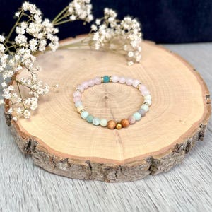 Può includere: Un braccialetto di perline rosa, blu e di legno, con dettagli dorati e turchesi. Il braccialetto è esposto su una fetta di legno, con fiori bianchi. Le perline misurano circa 0,8 cm di diametro.