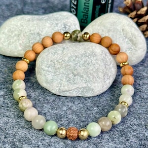Può includere: Un braccialetto di perline con perline di legno marrone chiaro, perline color oro e perline di pietra verde e bianca. Il braccialetto è appoggiato su una superficie grigia, con una bottiglia di olio essenziale sullo sfondo. La bottiglia ha la parola "Balance".