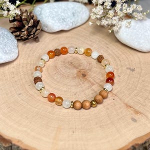 Könnte beinhalten: Ein Armband mit einer Mischung aus orangefarbenen, weißen und braunen Perlen, die durch kleine goldfarbene Abstandshalter unterbrochen werden. Das Armband besteht aus verschiedenen Naturstein- und Holzperlen. Das Armband wird auf einer Holzoberfläche präsentiert.